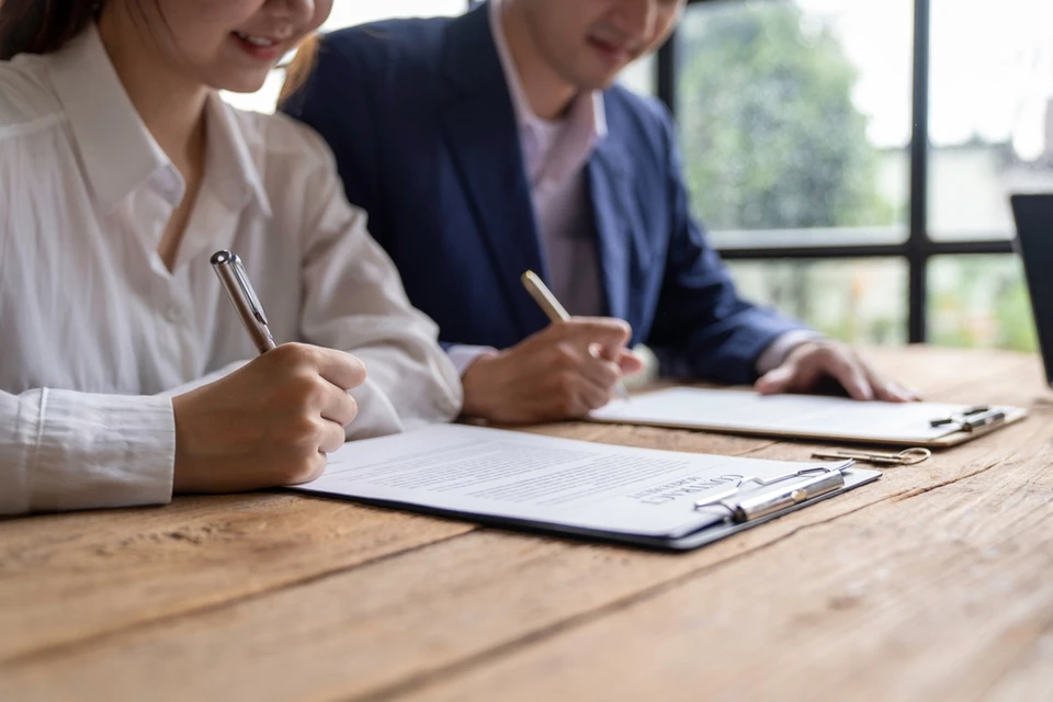 Deux professionnels en tenue d'affaires signent des documents contractuels dans un bureau moderne avec baie vitrée